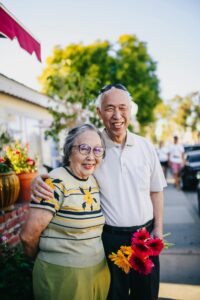 An elderly couple stands outside on a sunny day.